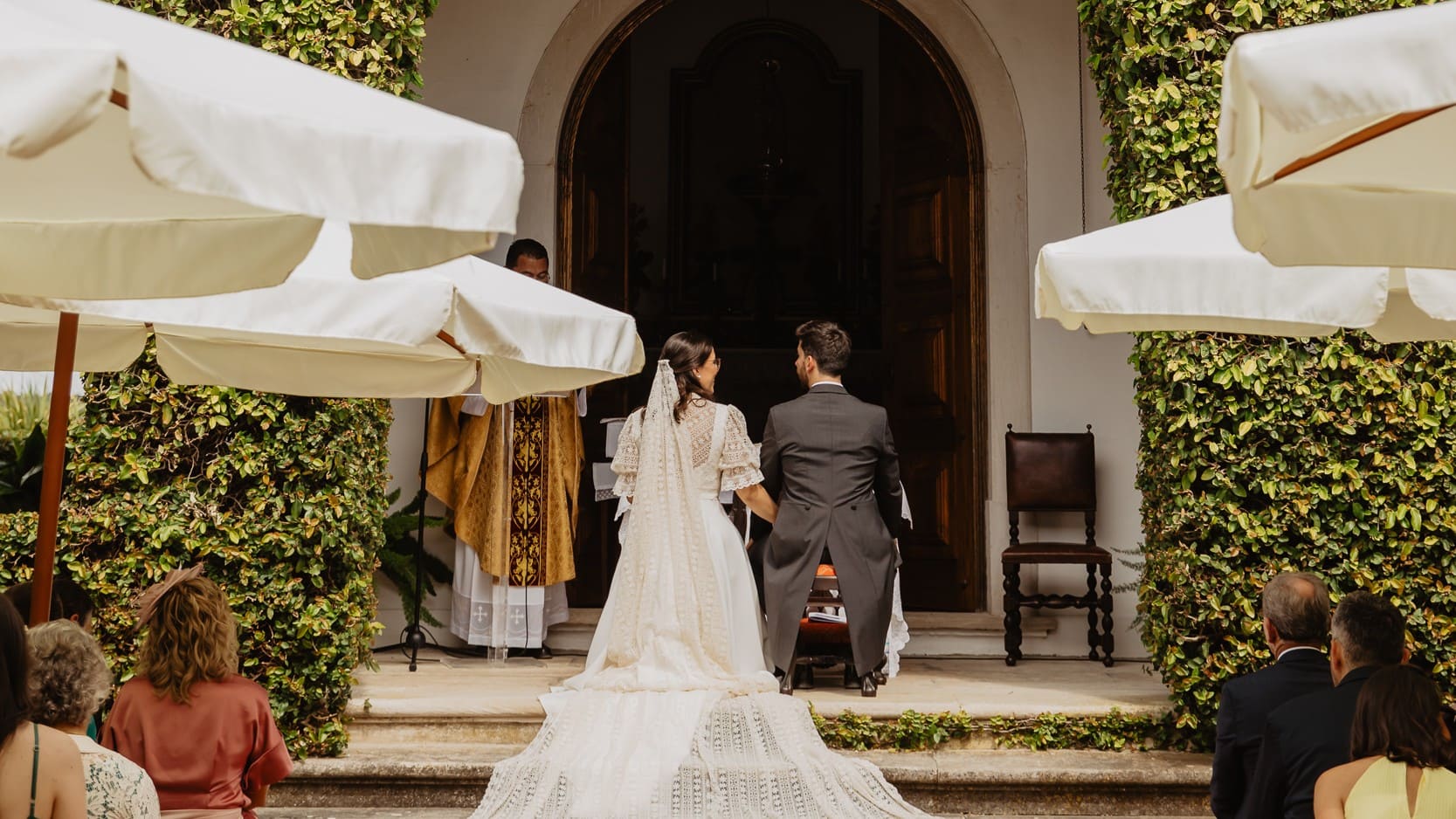 Cerimónia de casamento ao ar livre, com os noivos de costas em frente à capela
